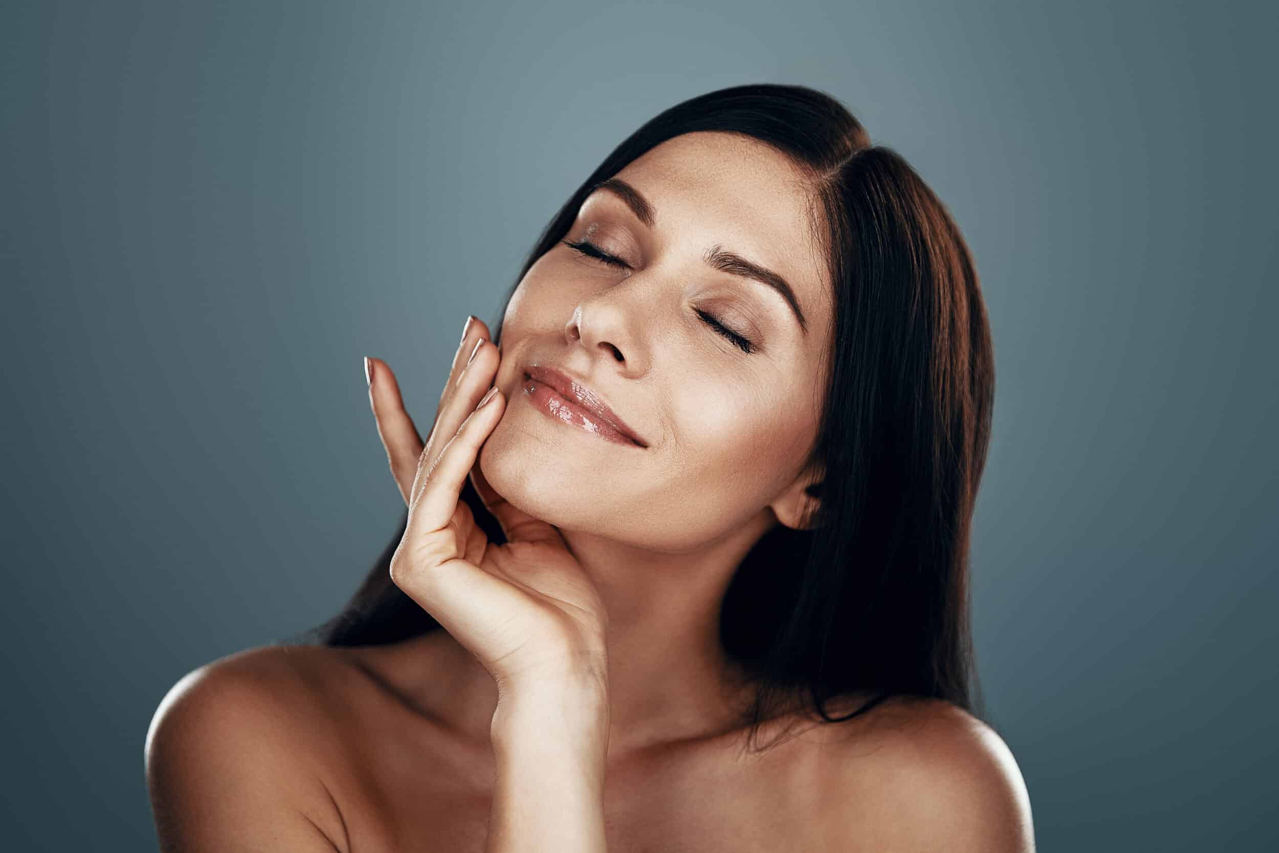 Beautiful young smiling woman touching her face and keeping eyes closed Beautiful young smiling woman touching her face and keeping eyes closed